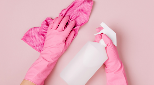 Pair of hands wearing pink rubber gloves, one holding a pink cloth, one holding white spray bottle on pink background